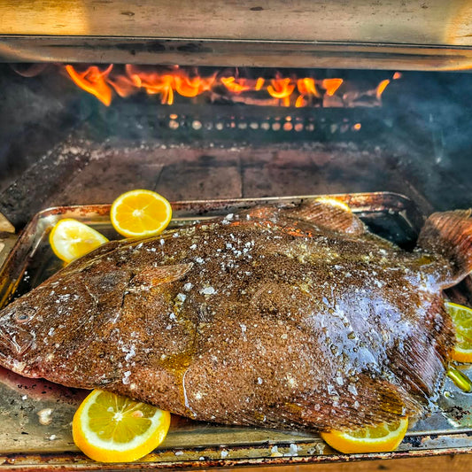 A whole grilled brill on a metal tray, seasoned with coarse salt and surrounded by lemon slices, set in an open barbecue grill with visible flames.