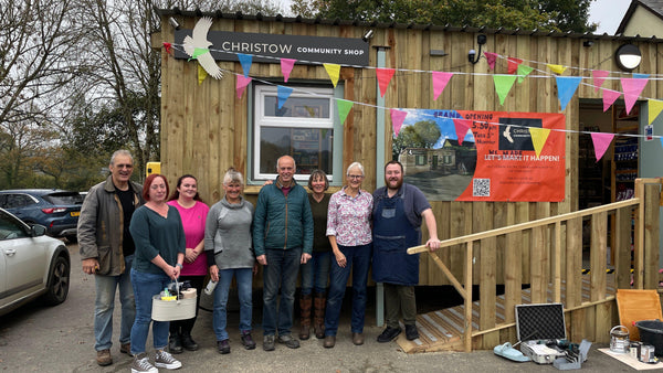 Group of people standing in front of a community shop with bunting and a sign.