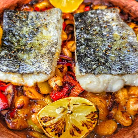 A close-up shot of a prepared fish dish in a clay pot, featuring two grilled fish fillets on top of a bed of chickpeas, red peppers, and lemon slices.