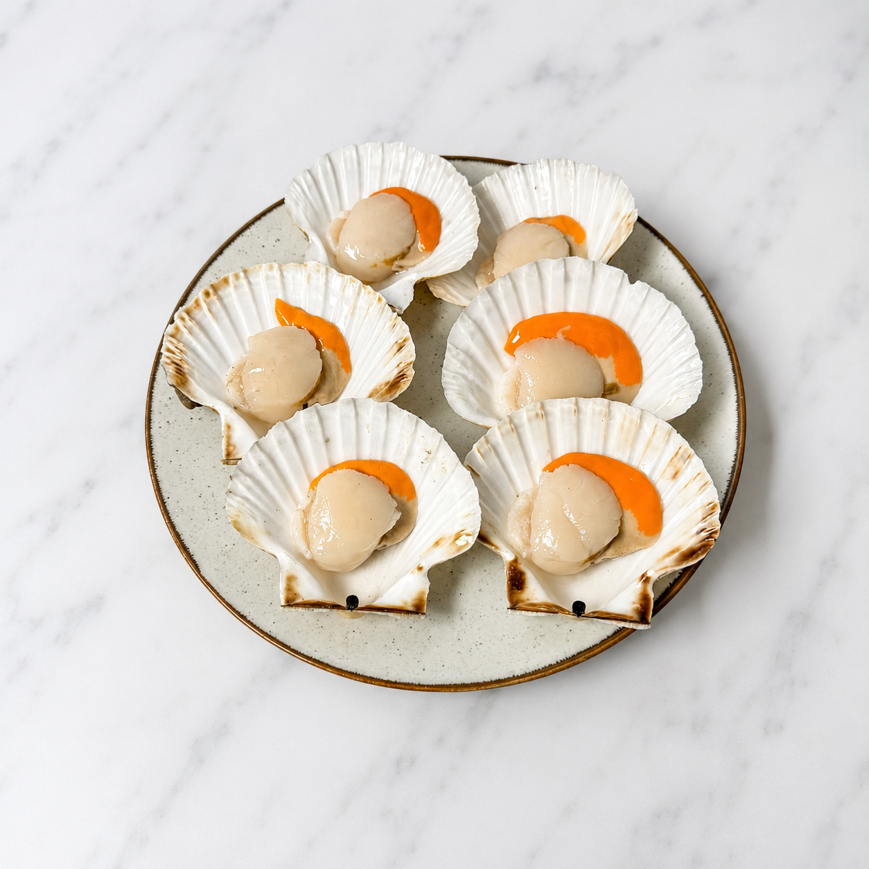 Six cooked scallops presented in white scallop shells with orange sauce, arranged on a speckled ceramic plate over a white marble surface.