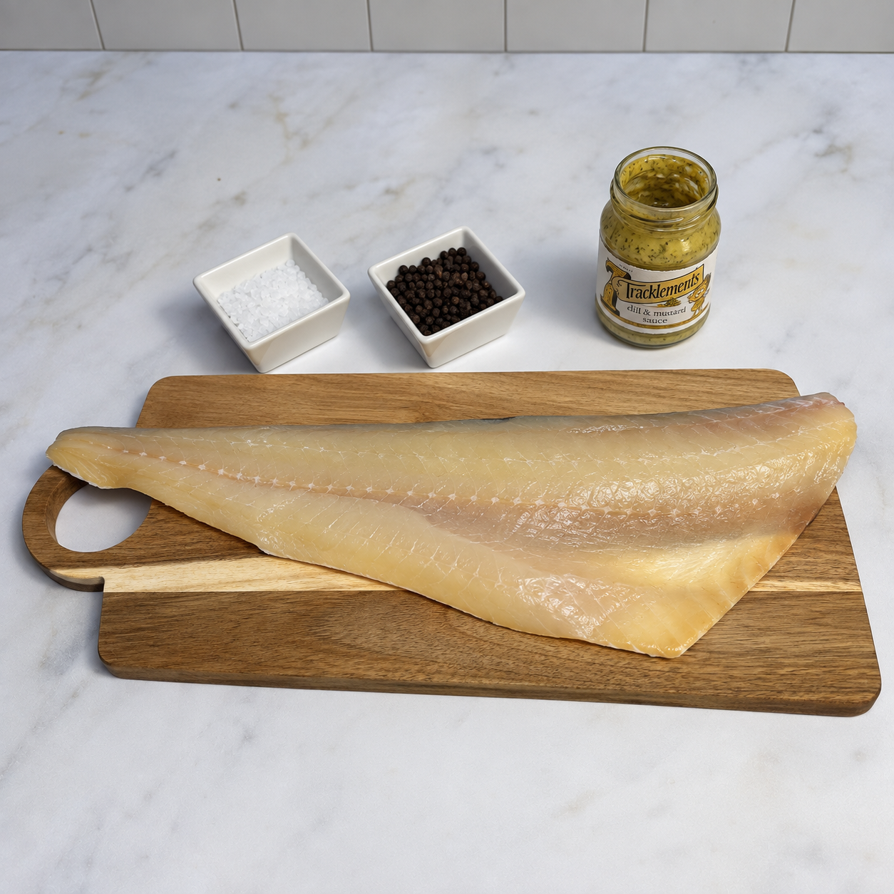 A large raw fillet of smoked haddock resting on a wooden cutting board on a white marble countertop, with two small white dishes containing salt and peppercorns and an open jar of Tracklements dill and mustard sauce behind it.