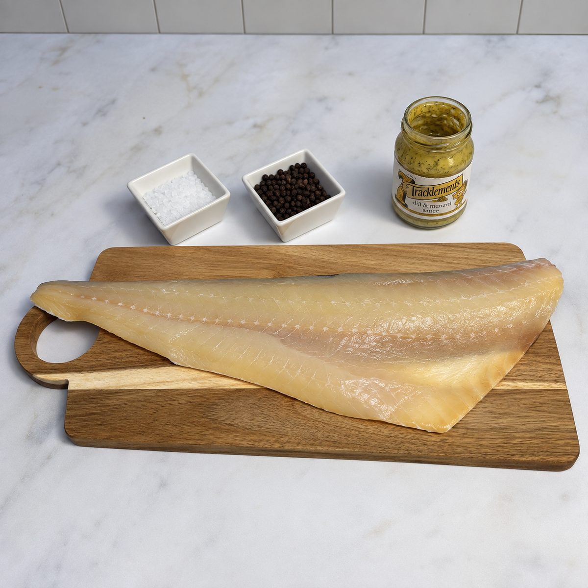 A large raw fillet of smoked haddock resting on a wooden cutting board on a white marble countertop, with two small white dishes containing salt and peppercorns and an open jar of Tracklements dill and mustard sauce behind it.